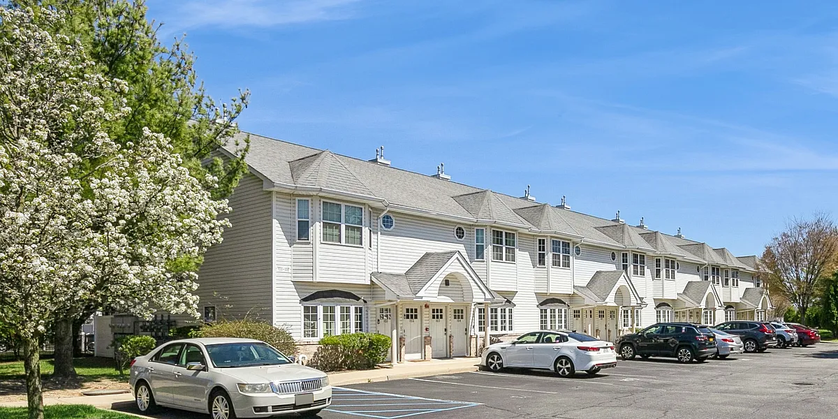 Charming townhouse complex with spacious parking, surrounded by lush greenery and blossoming trees under a vibrant blue sky.
