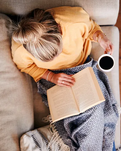 Cozy living space featuring a person relaxing on a sofa with a book and coffee, adjacent to a clean, organized table with plants and reading materials.