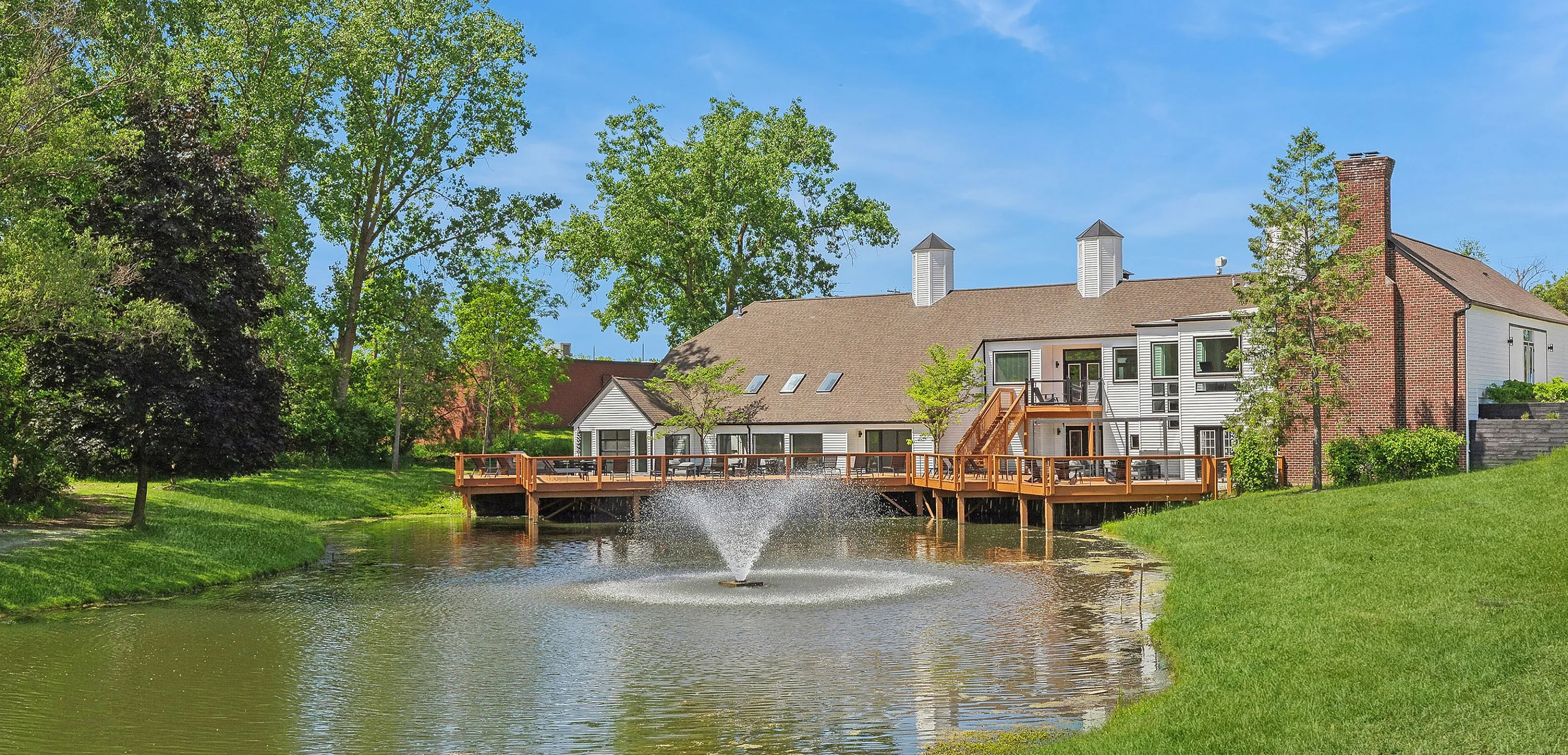 Charming apartment complex with scenic pond and fountain, surrounded by lush greenery and bright blue skies.