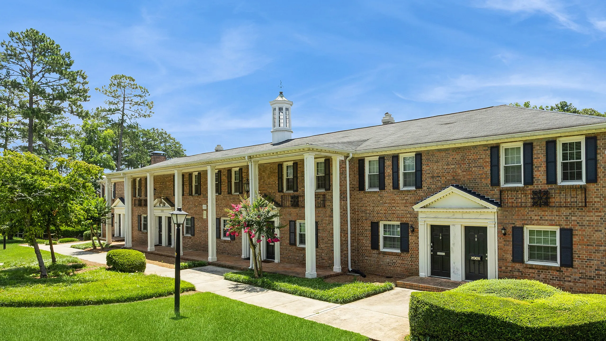 Charming brick apartment complex with colonial-style architecture, lush greenery, and a bright, sunny sky.