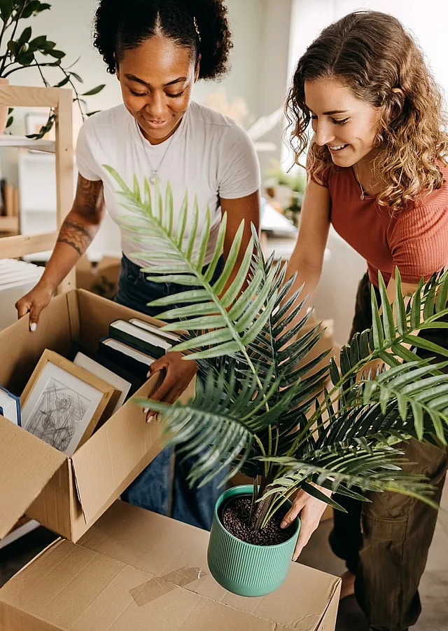 Joyful home move-in scene with two people unpacking boxes in a bright, plant-filled room.