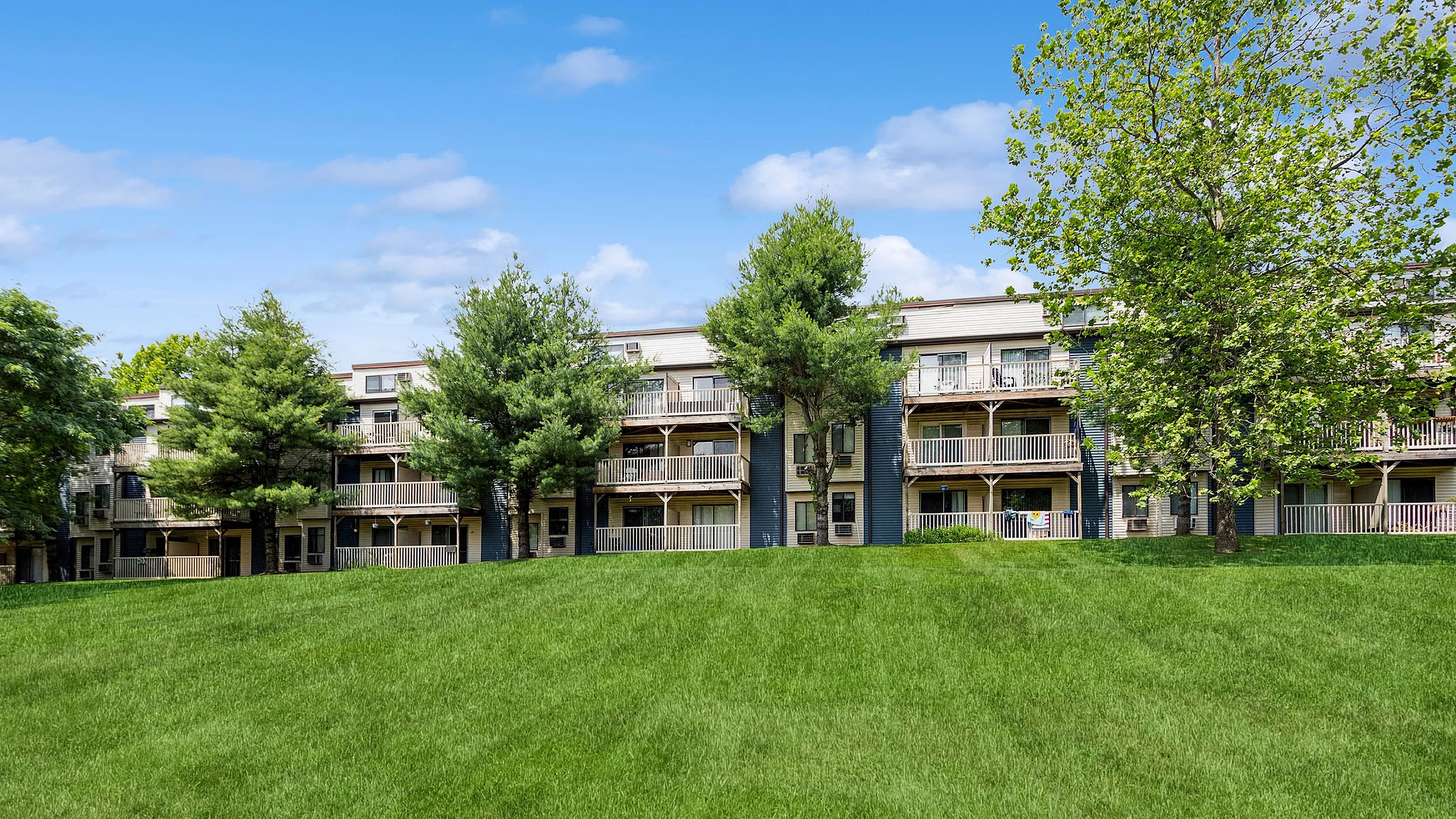 Charming apartment complex with lush green lawns and spacious balconies, nestled among mature trees under a bright blue sky.