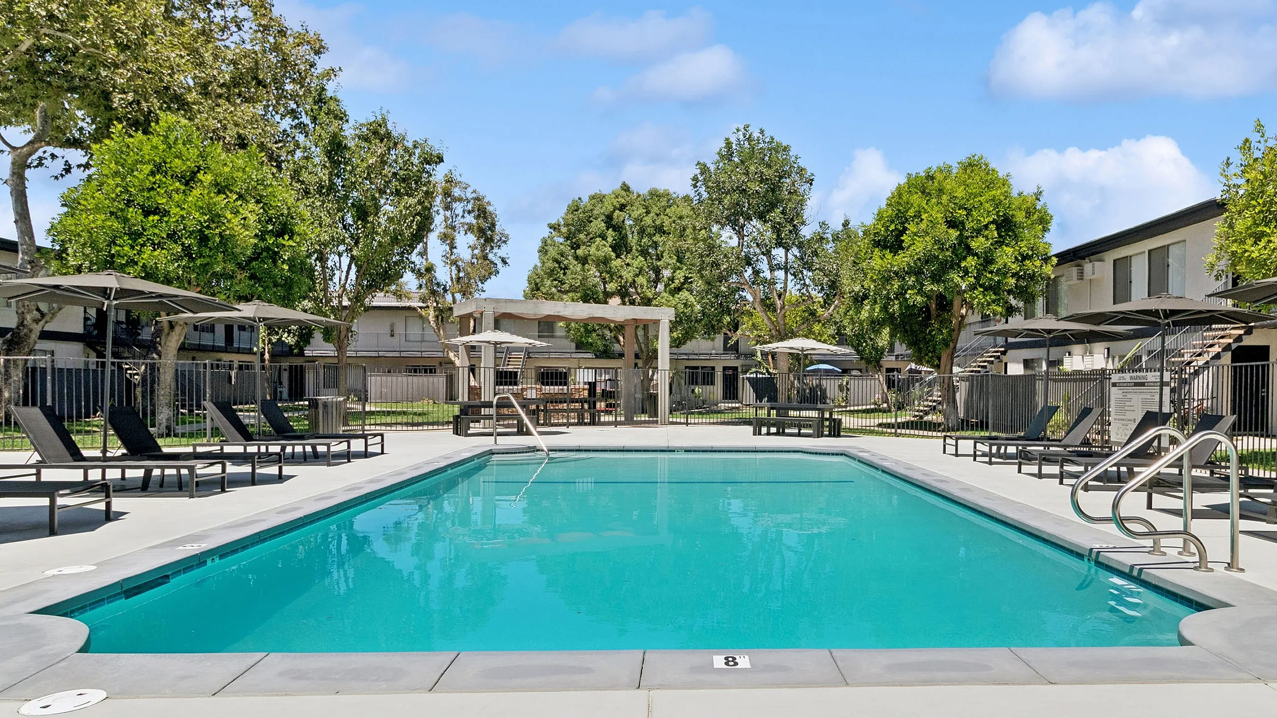 Inviting outdoor swimming pool with sleek lounge chairs and umbrellas, surrounded by lush greenery and stylish apartment buildings.