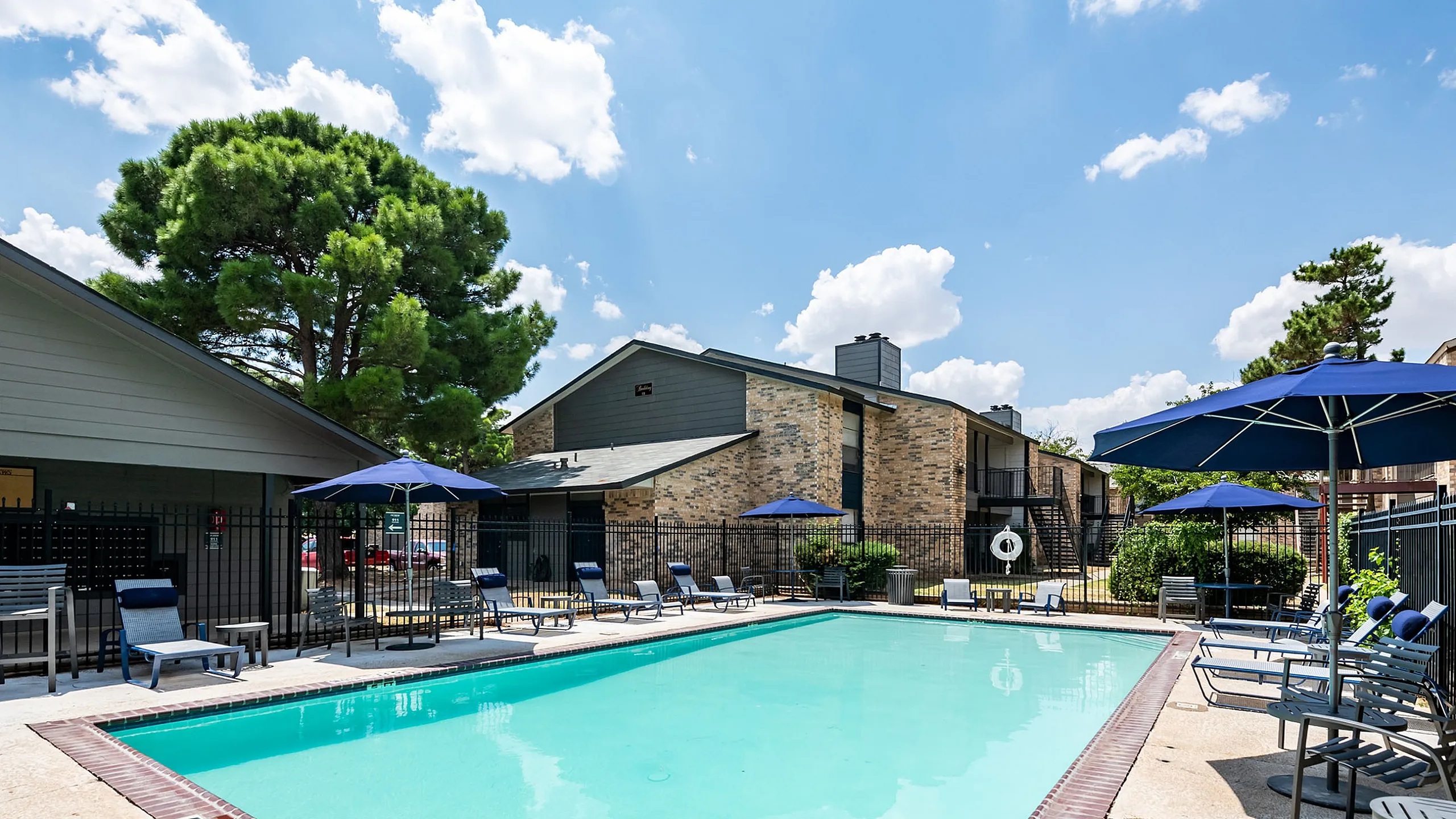Inviting apartment pool area with sun loungers, blue umbrellas, and lush surroundings, perfect for relaxation and enjoyment.