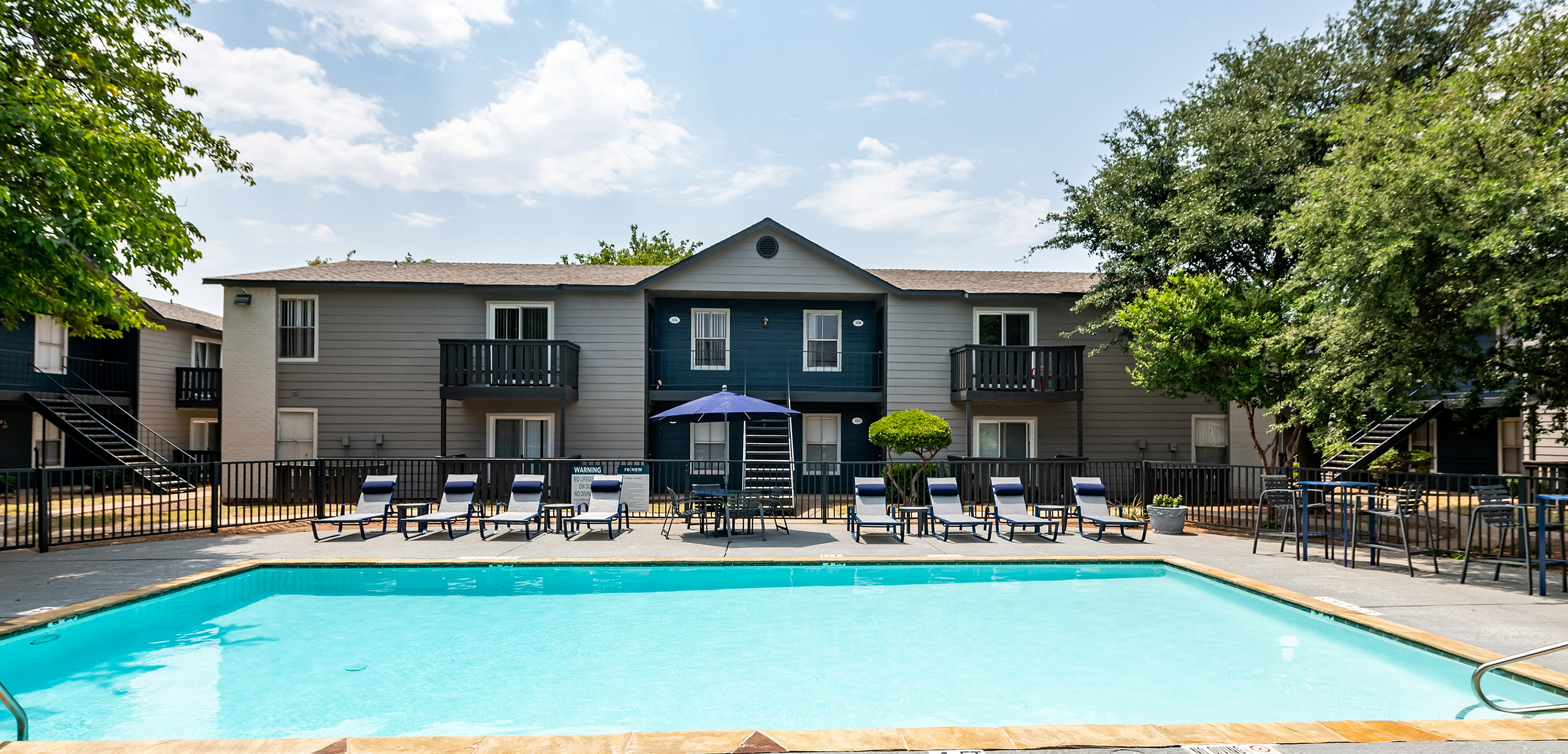Inviting apartment complex with sparkling outdoor pool, surrounded by comfortable lounge chairs and lush greenery, under a clear blue sky.