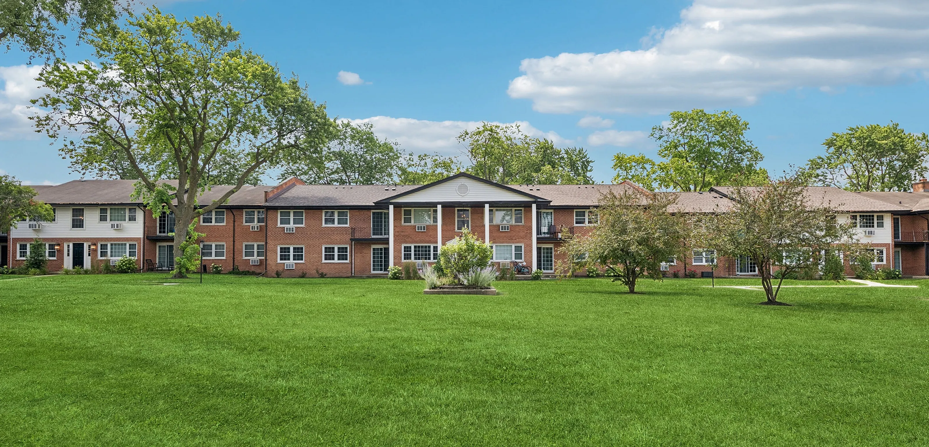 Charming brick apartment complex surrounded by lush greenery and vibrant trees, under a bright blue sky.