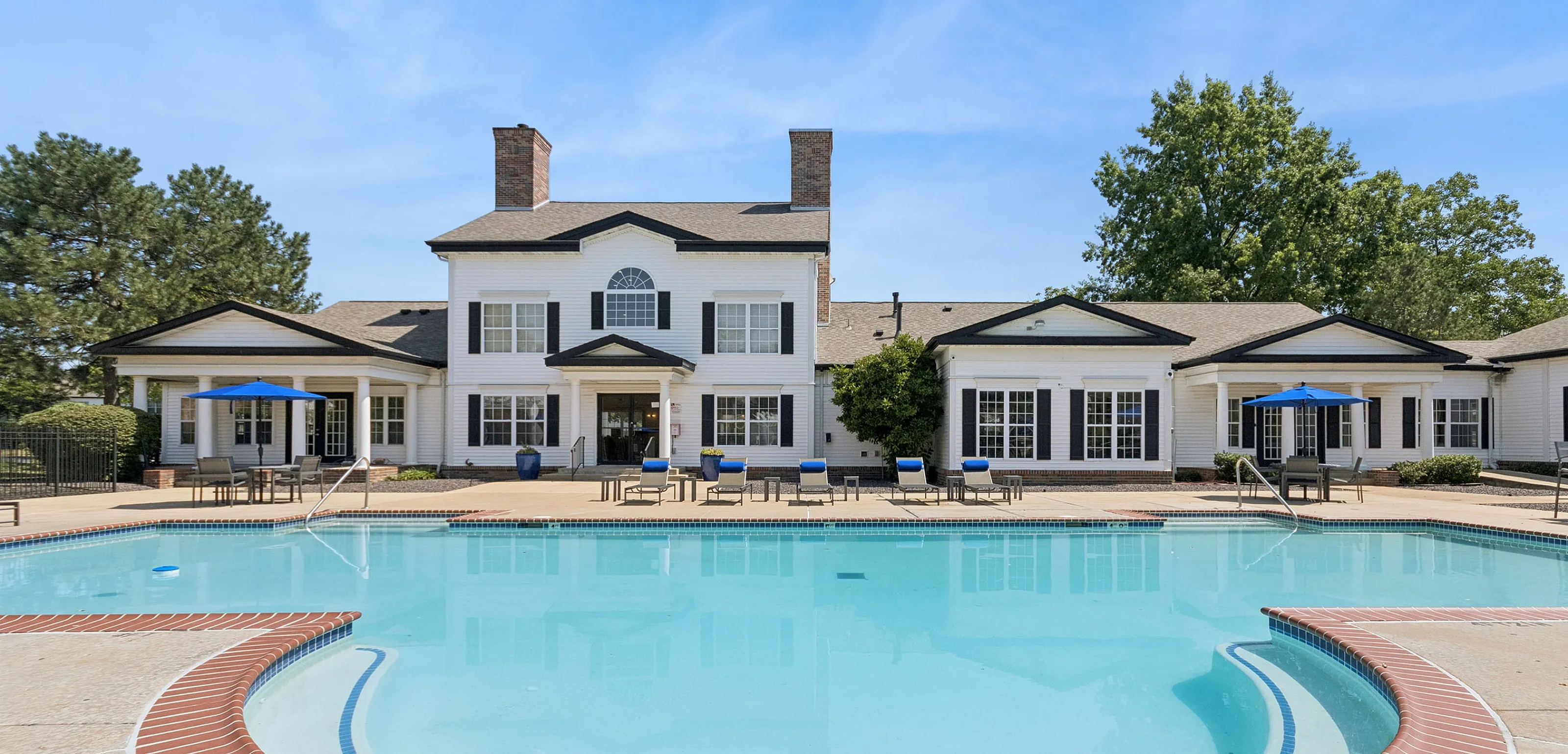 Inviting pool area in front of a charming suburban clubhouse, featuring elegant white architecture and lush greenery, perfect for relaxation and leisure.