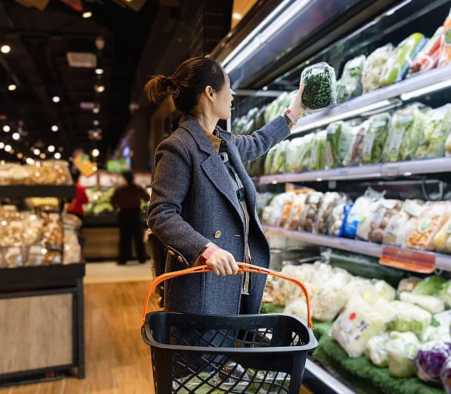 A shopper in a stylish coat selects fresh greens from a well-stocked produce aisle, highlighting a vibrant and inviting grocery shopping experience.