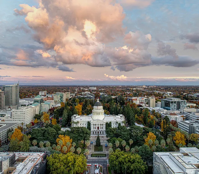 Breathtaking aerial view of a vibrant cityscape highlighting lush greenery, a majestic classical building, and dramatic sunset clouds.