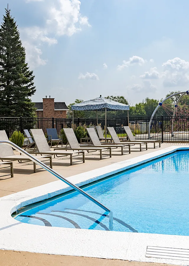 Inviting outdoor pool area with sun loungers, surrounded by greenery and a modern playground in the background, perfect for relaxation and recreation.