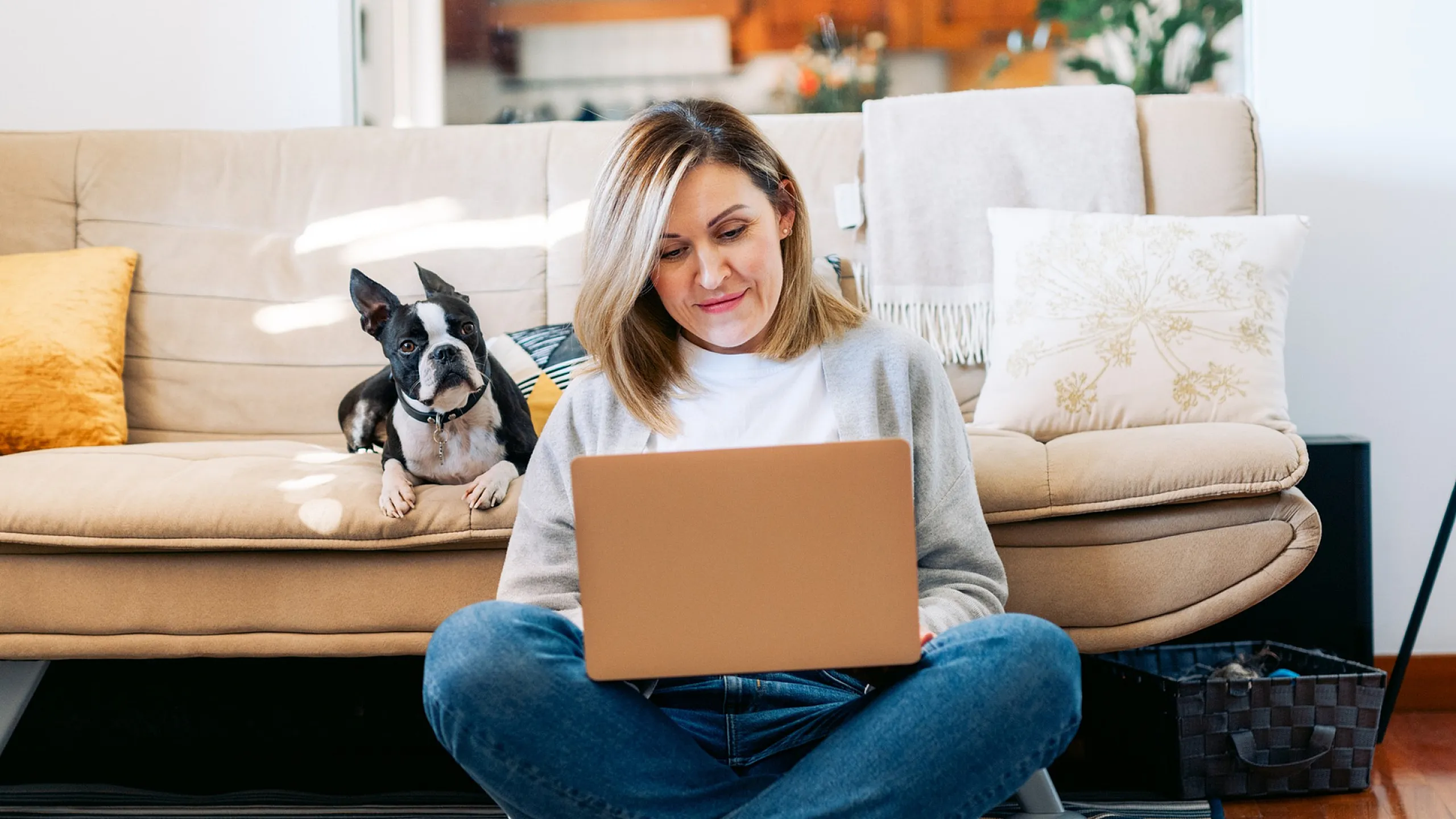 Cozy living room scene with a woman working on a laptop, accompanied by a cute dog on the sofa, surrounded by soft cushions and warm lighting.