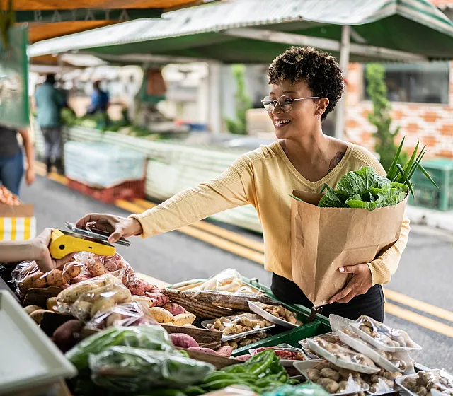 Vibrant farmers' market scene with a smiling shopper holding a bag of fresh greens, making a purchase of assorted produce under a green canopy.