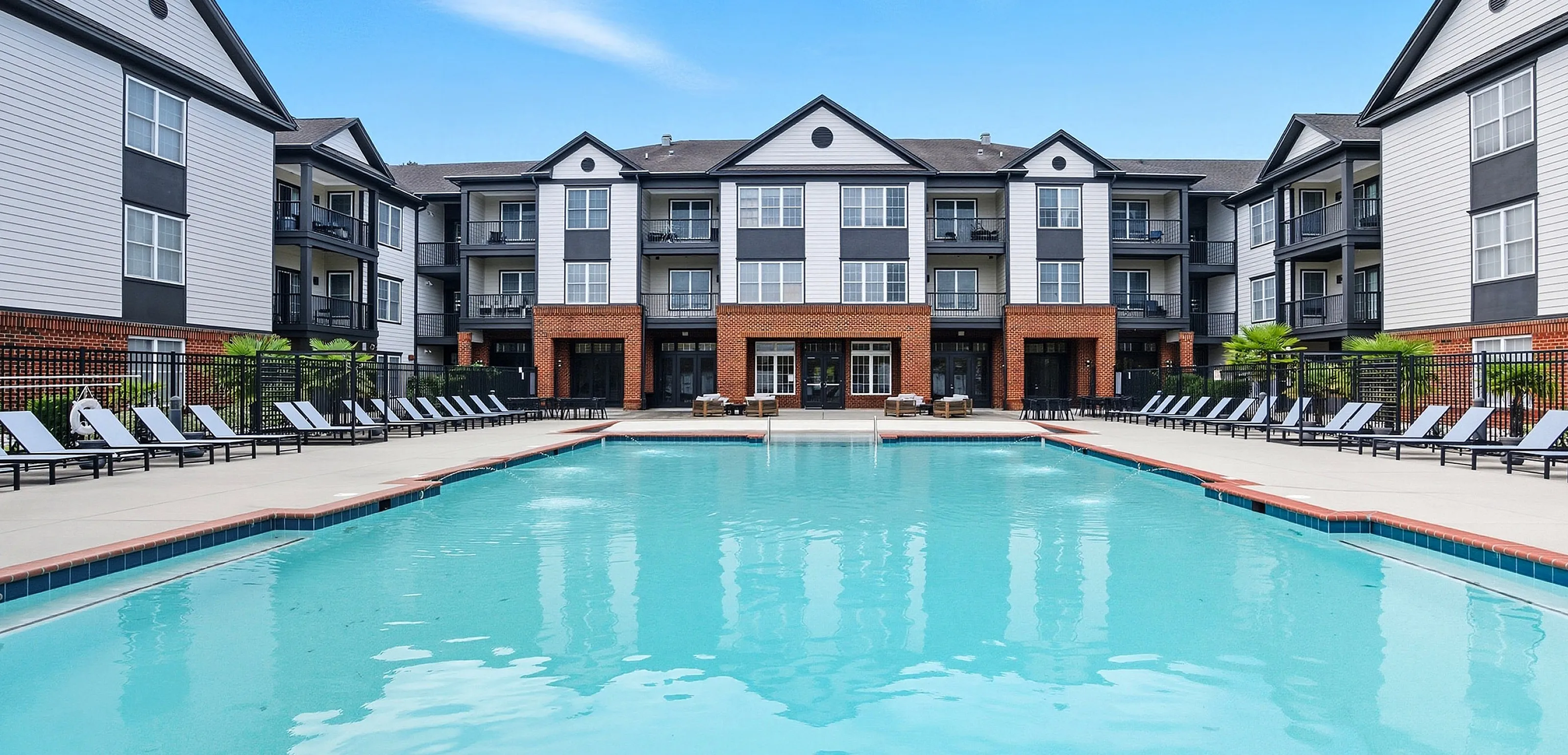 Luxury apartment complex with a sparkling blue swimming pool, surrounded by modern architecture and stylish lounge chairs, under a bright blue sky.