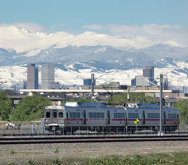 Scenic train ride passing through lush greenery with a breathtaking view of snow-capped mountains and a bustling city skyline.