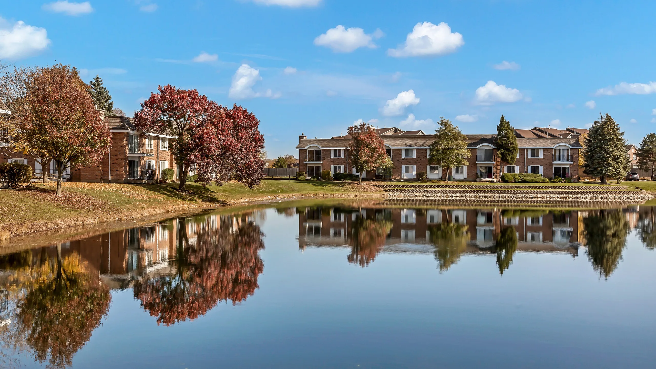 Serene apartment complex with charming brick facade, nestled by a tranquil lake and vibrant autumn trees reflecting in the water.