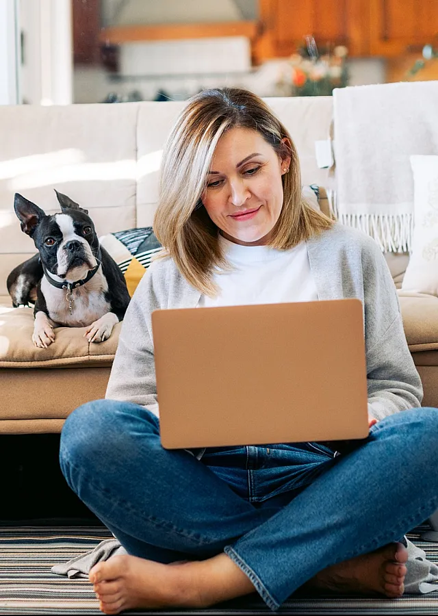 Cozy living room with inviting beige sofa, cheerful woman working on a laptop, and a playful dog in a spacious, well-lit apartment.