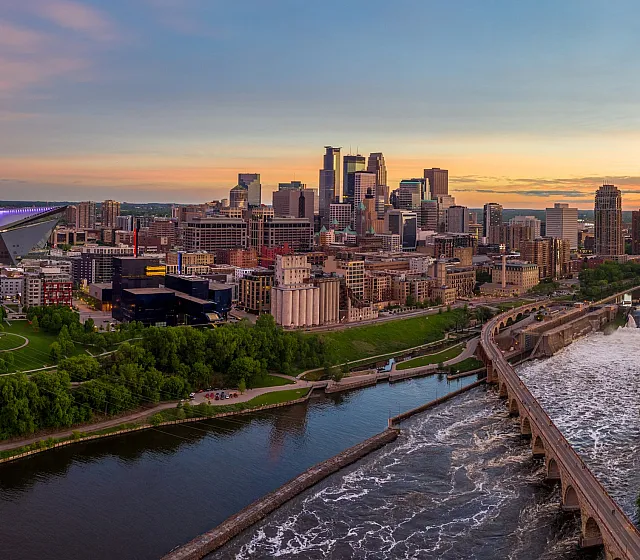 Captivating panoramic view of a vibrant Minneapolis city skyline at sunset, featuring modern skyscrapers, a prominent river, lush green parks, and an iconic stadium with brilliant lighting.