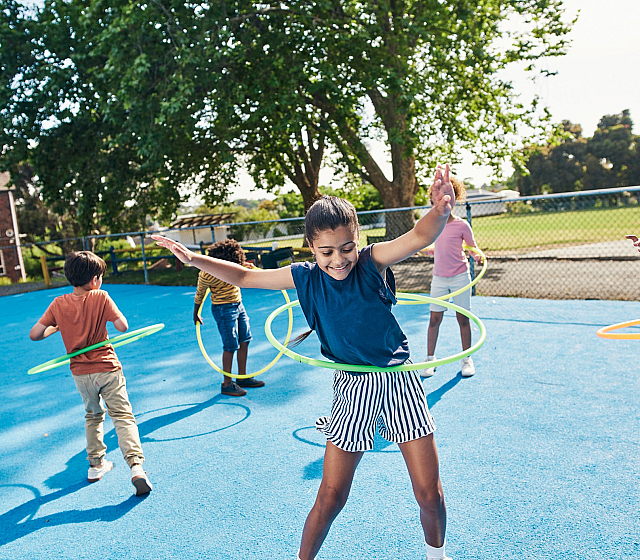 Children enjoying outdoor play with hula hoops on a vibrant blue playground, surrounded by lush greenery.