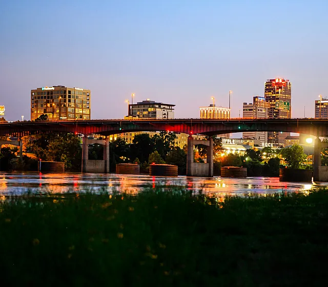 Stunning Little Rock cityscape view at sunset showcasing illuminated skyscrapers, a reflective water bridge, and vibrant urban lights.