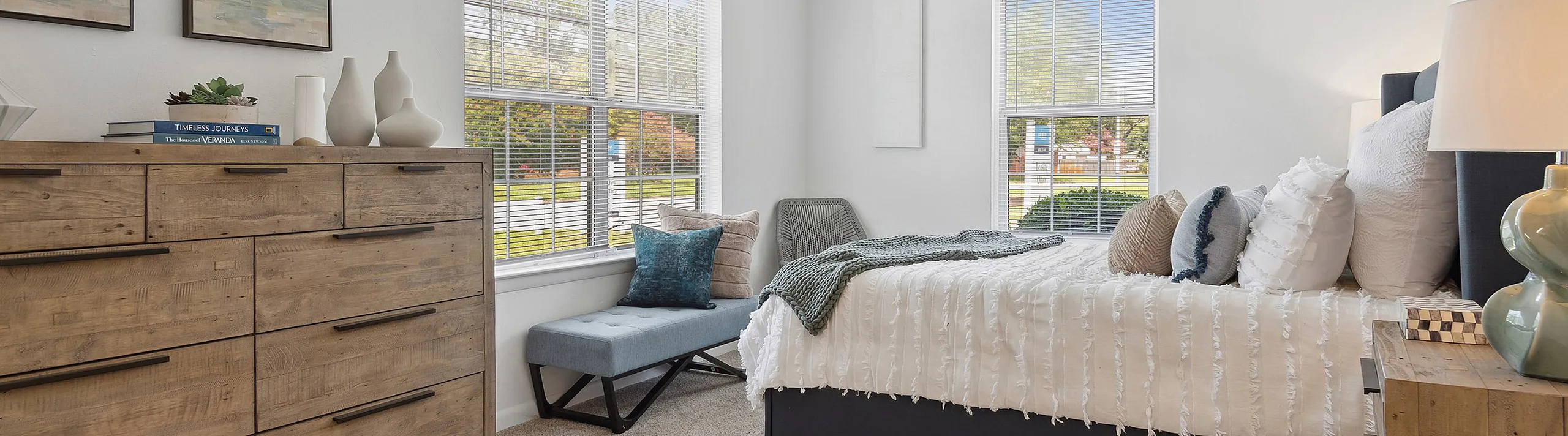 Inviting bedroom at ReNew Virginia Beach with large windows, cozy bed, elegant decor, and a stylish wooden dresser.