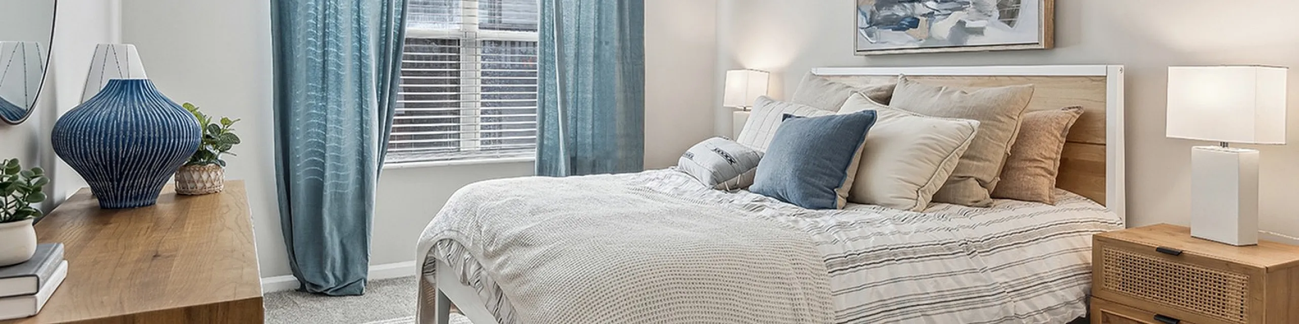 Elegant bedroom at ReNew Marina Shores featuring soft blue drapes, contemporary artwork, and a cozy bed with layered pillows, creating a serene and inviting ambiance.