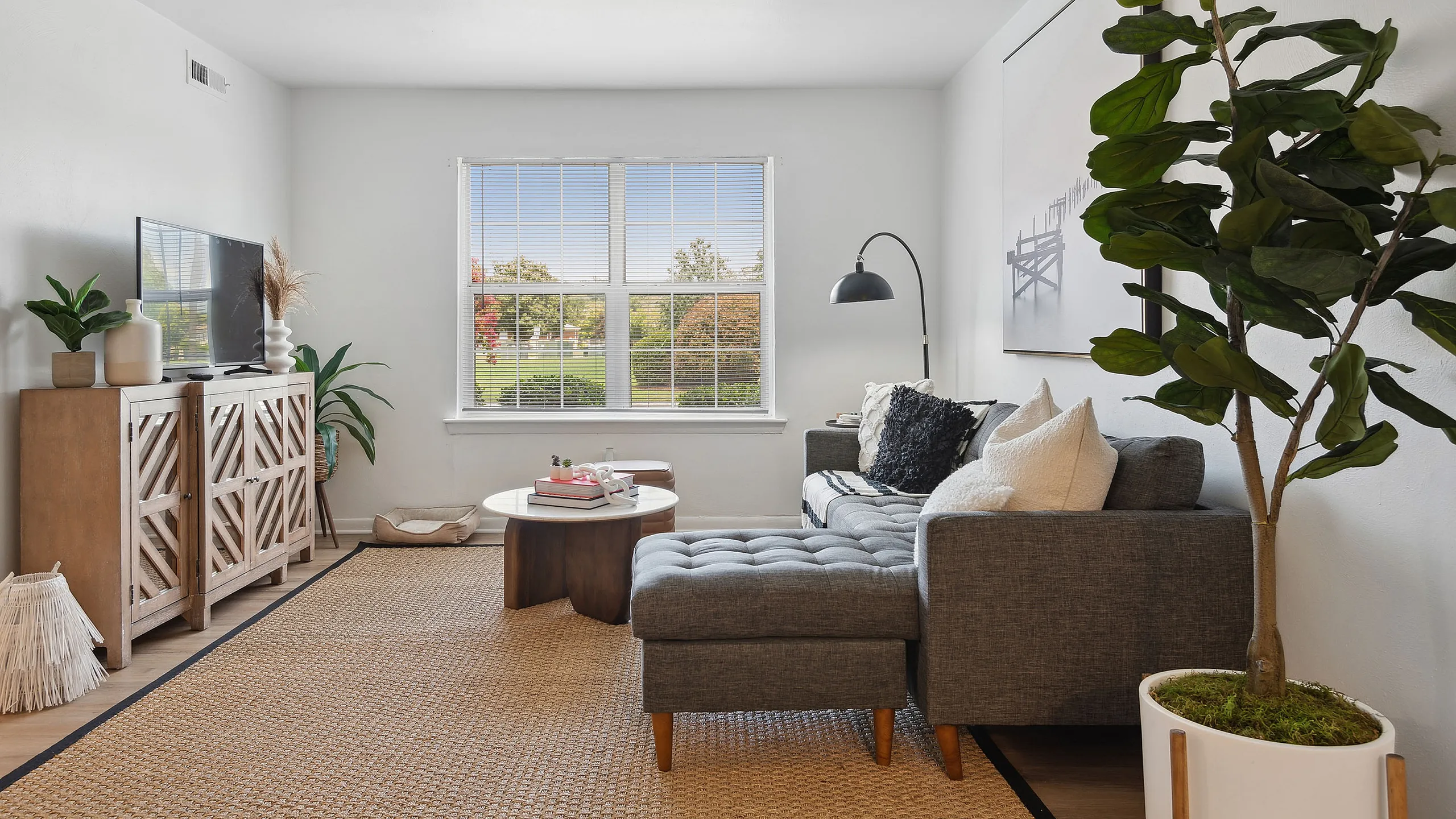 Bright and inviting living room at ReNew Virginia Beach featuring a cozy gray sectional, stylish decor, and a large window providing a scenic view of a lush green landscape.