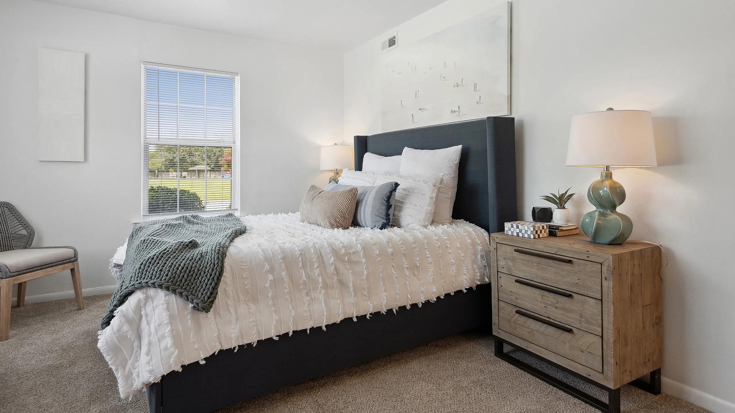Bright, inviting bedroom at ReNew Virginia Beach with stylish decor, featuring a cozy bed adorned with plush pillows and a soft throw blanket, complemented by a chic nightstand and modern lamp.