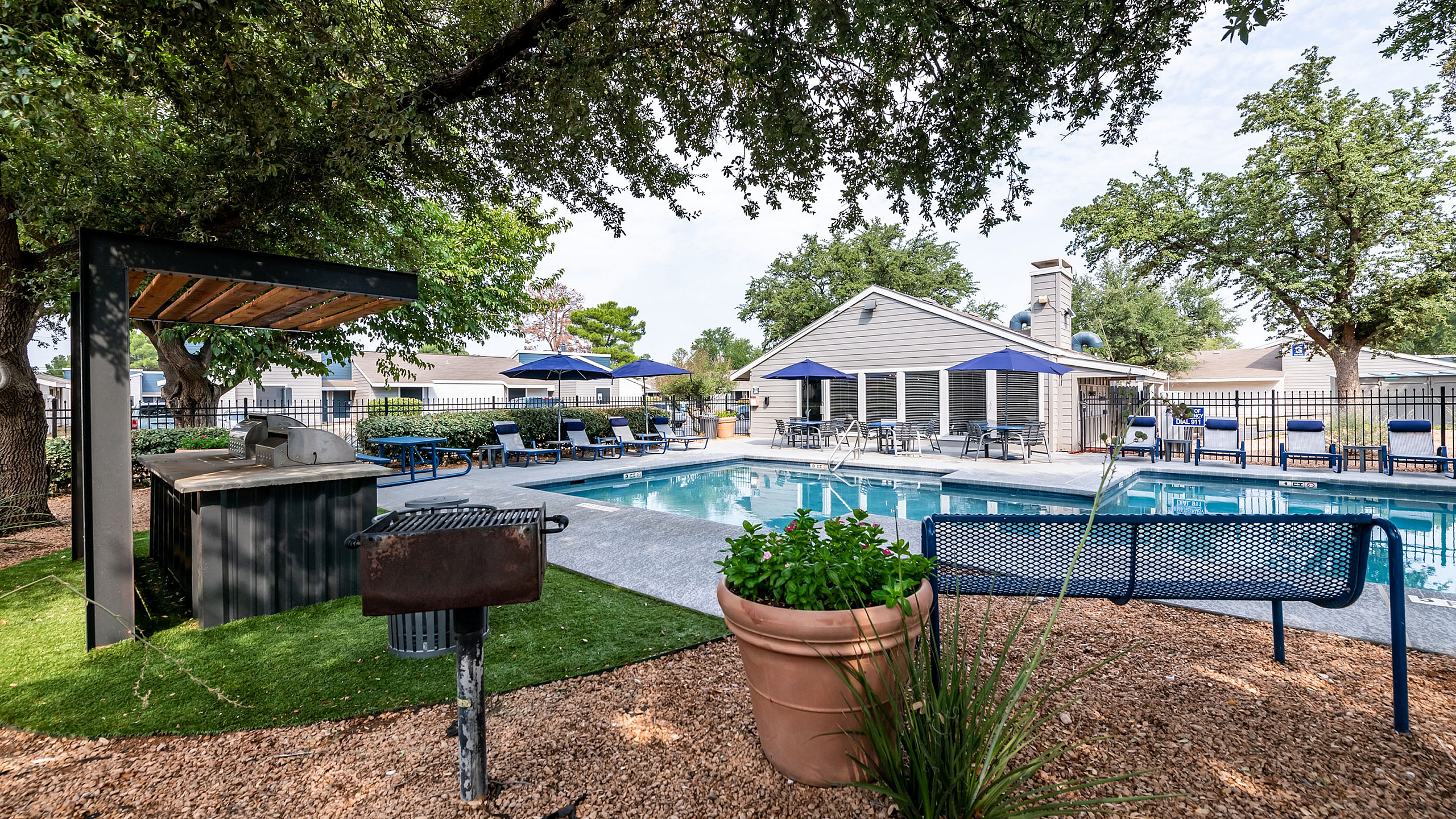 Charming outdoor area at ReNew Heights featuring a sparkling pool, ample lounge chairs with blue umbrellas, a barbecue grill, and greenery under shade trees.
