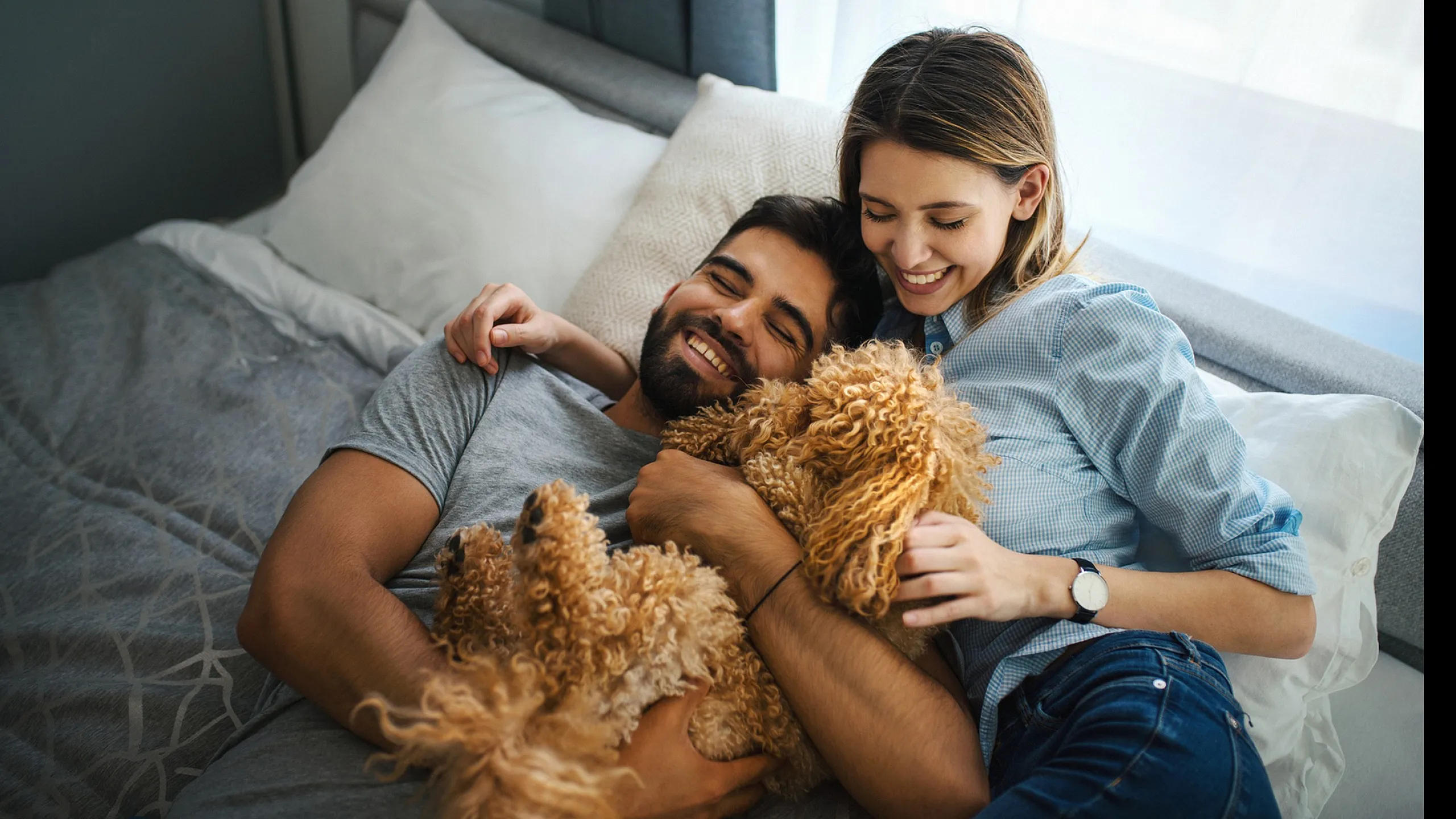 Joyful couple relaxing in a cozy bedroom with a fluffy dog, creating a warm and inviting atmosphere.