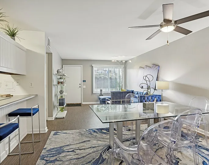 Bright and modern living space with open layout, sleek dining area with glass table, and stylish blue accents on furniture.