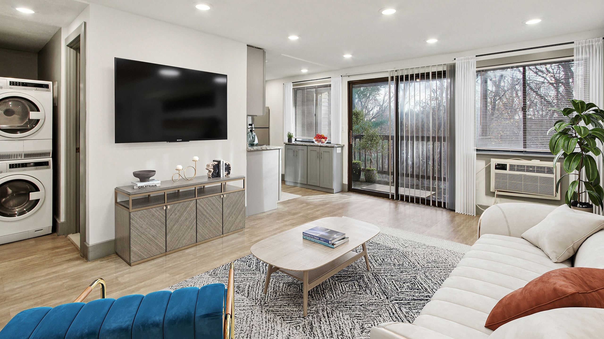 Modern, open-concept living space with elegant decor features a cozy sofa, vibrant blue armchair, and large wall-mounted TV. Sunlit room includes a stacked washer-dryer, sleek kitchen, and expansive windows with serene views of the outdoors