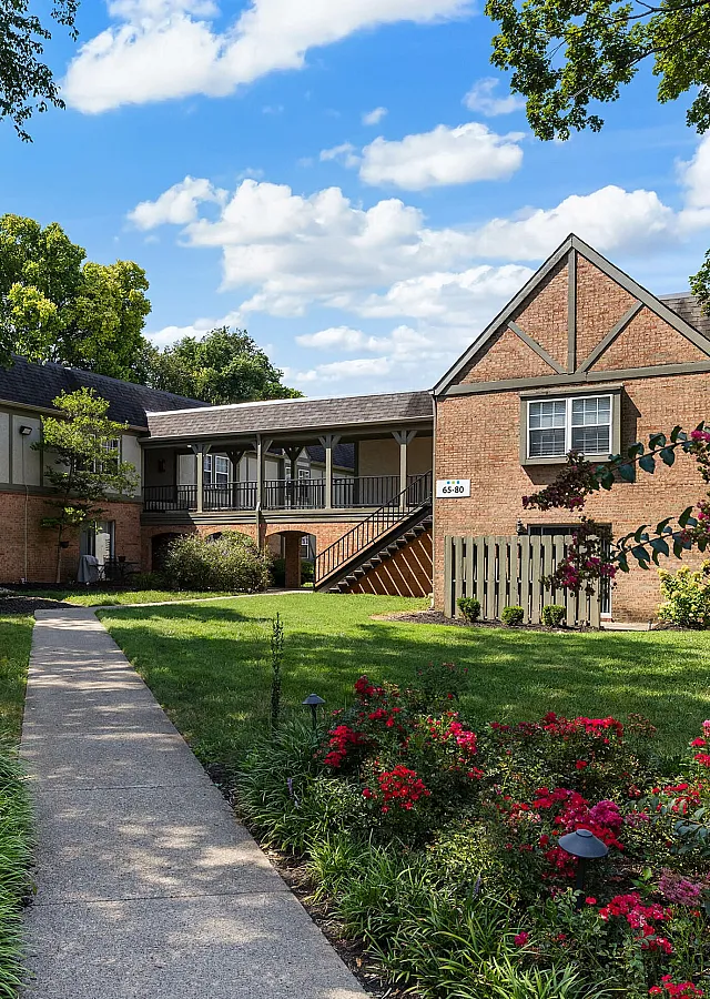 Charming apartment complex surrounded by lush greenery and vibrant flowers, featuring a classic brick exterior and a welcoming pathway.