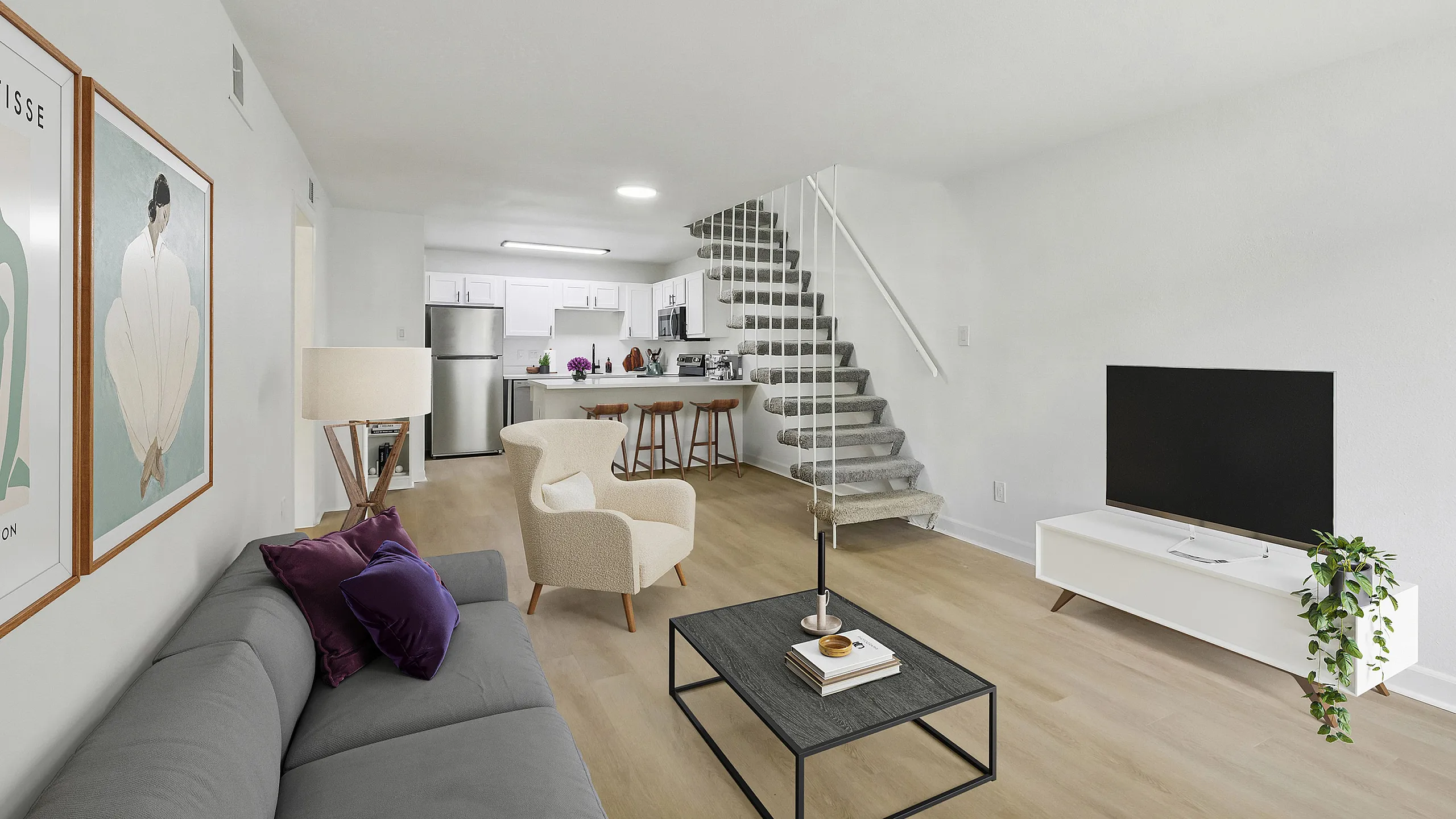 Modern living room with stylish decor, featuring a cozy gray couch, elegant artwork, open-concept kitchen with bar stools, and sleek staircase enhancing a spacious layout.