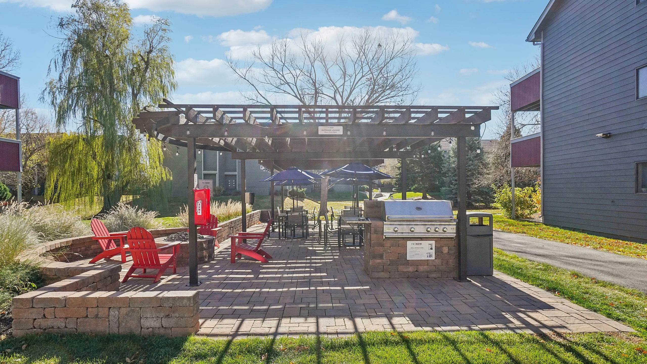 Charming outdoor grilling area with a pergola, surrounded by lush greenery and vibrant red chairs, ideal for entertaining and relaxation.