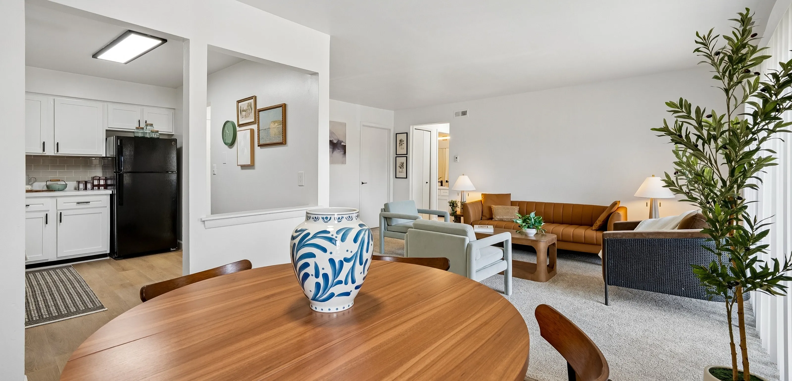 Elegant open-plan living space with a cozy seating area, stylish wooden dining table, and modern kitchen featuring white cabinetry and a sleek black refrigerator.
