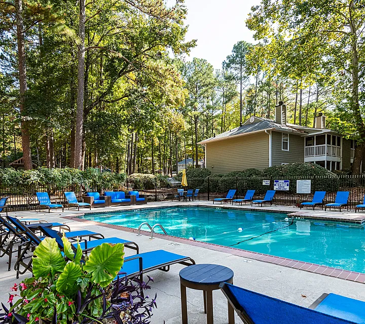 Tranquil apartment complex pool area surrounded by lush greenery, featuring stylish blue lounge chairs and inviting seating, perfect for relaxation and leisure.