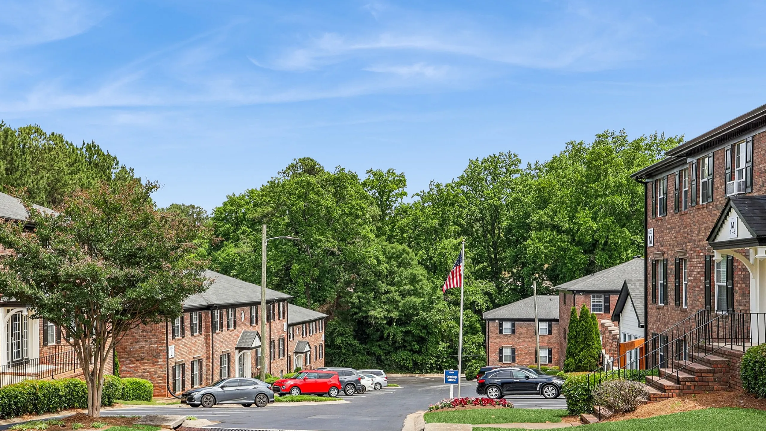 Charming apartment complex with brick buildings, lush greenery, and ample parking on a sunny day.
