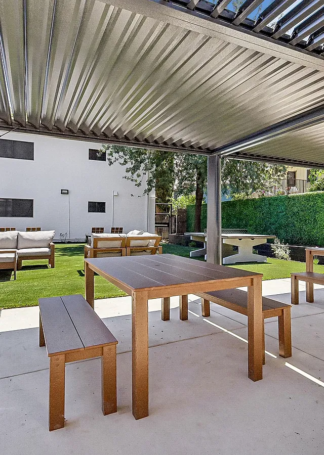 Charming outdoor seating area under a pergola with wooden tables and benches, surrounded by lush greenery and a manicured lawn.