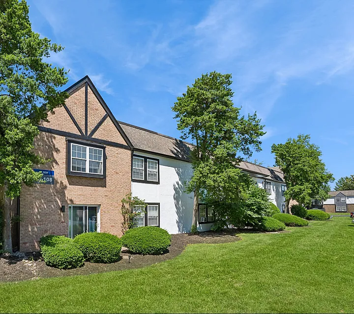 Charming apartment building with classic brick and white exterior, surrounded by lush green landscaping and tall trees.