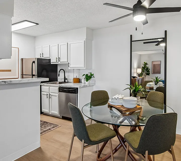 Modern apartment kitchen and dining area featuring sleek white cabinets, stainless steel appliances, and a stylish glass-top table with green chairs, illuminated by a chic ceiling fan.