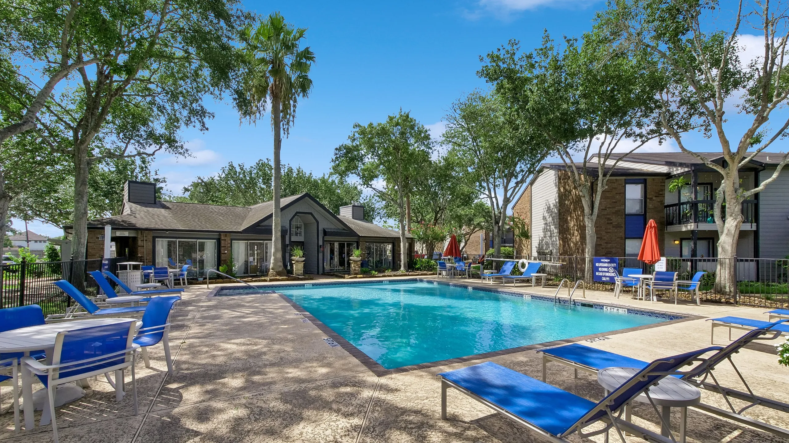 Inviting apartment complex pool area with lush landscaping, modern loungers, and shaded seating under a clear blue sky.