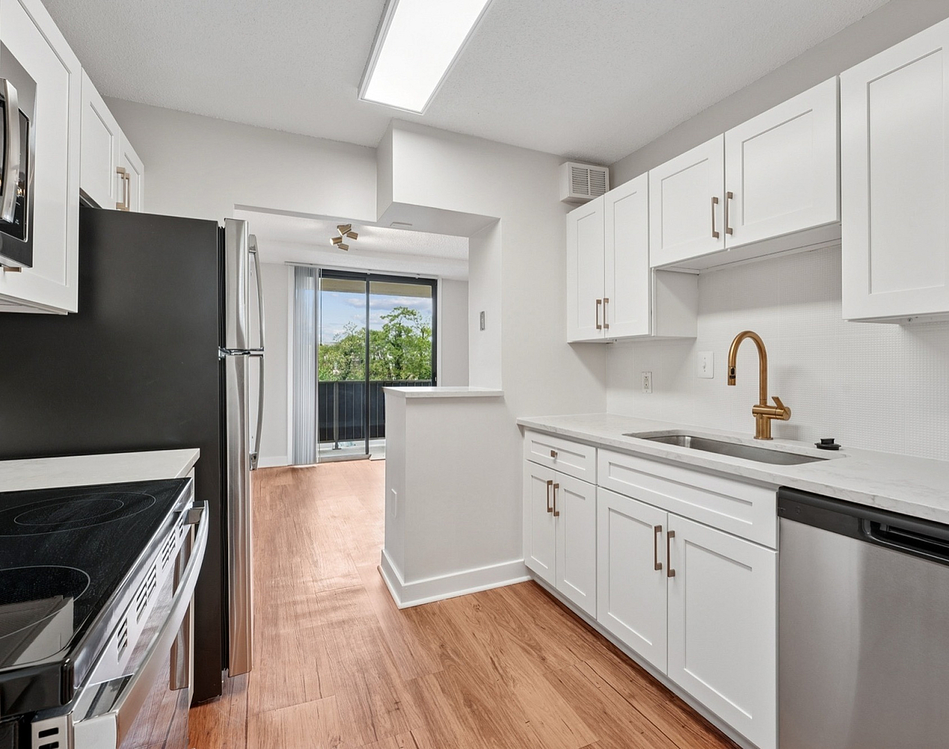 Modern kitchen with sleek white cabinetry, stainless steel appliances, and elegant wood flooring, leading to a bright living area with a scenic view.