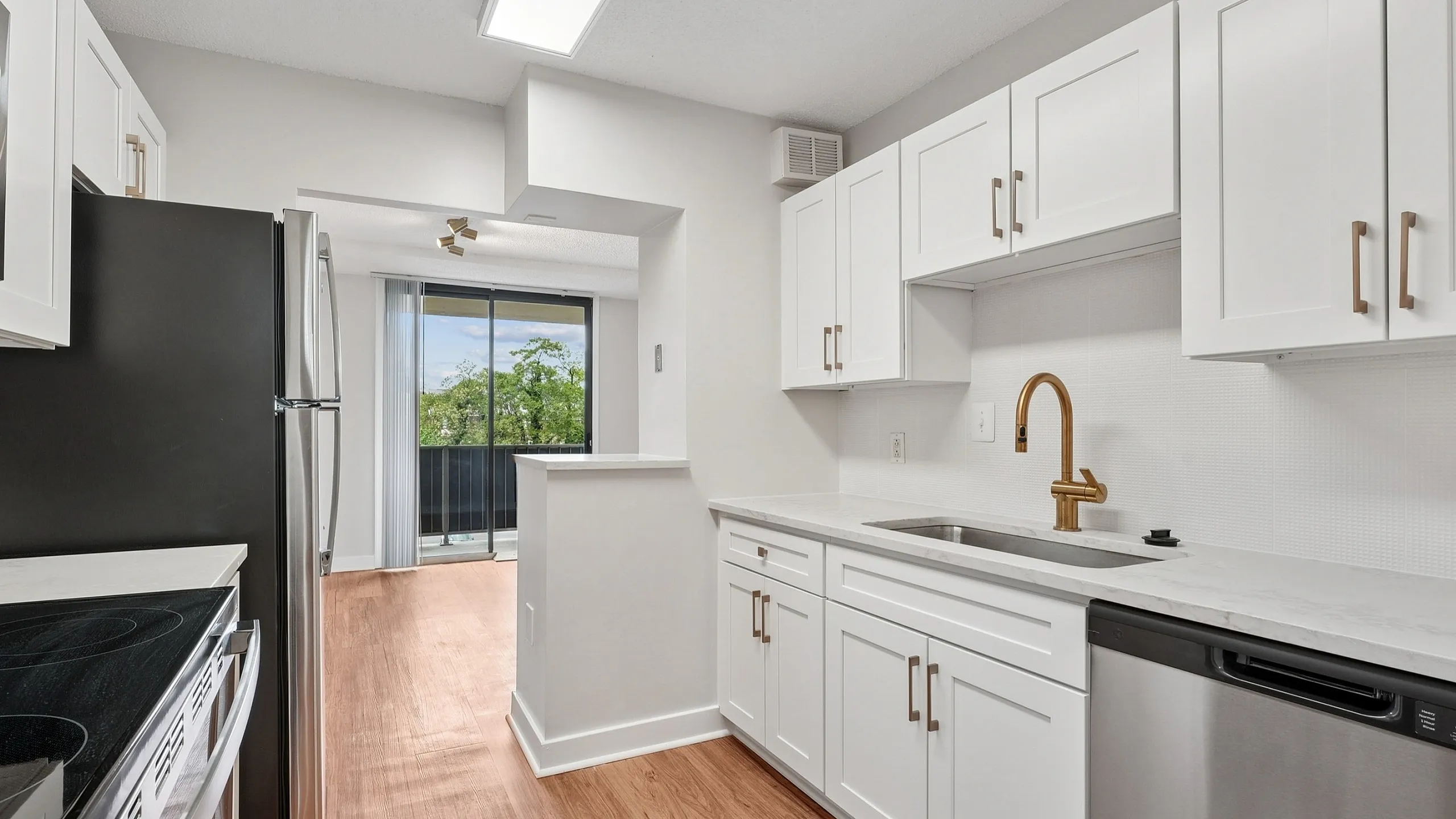 Contemporary kitchen with sleek stainless steel appliances, white cabinets, and elegant wood flooring leading to a bright balcony view.