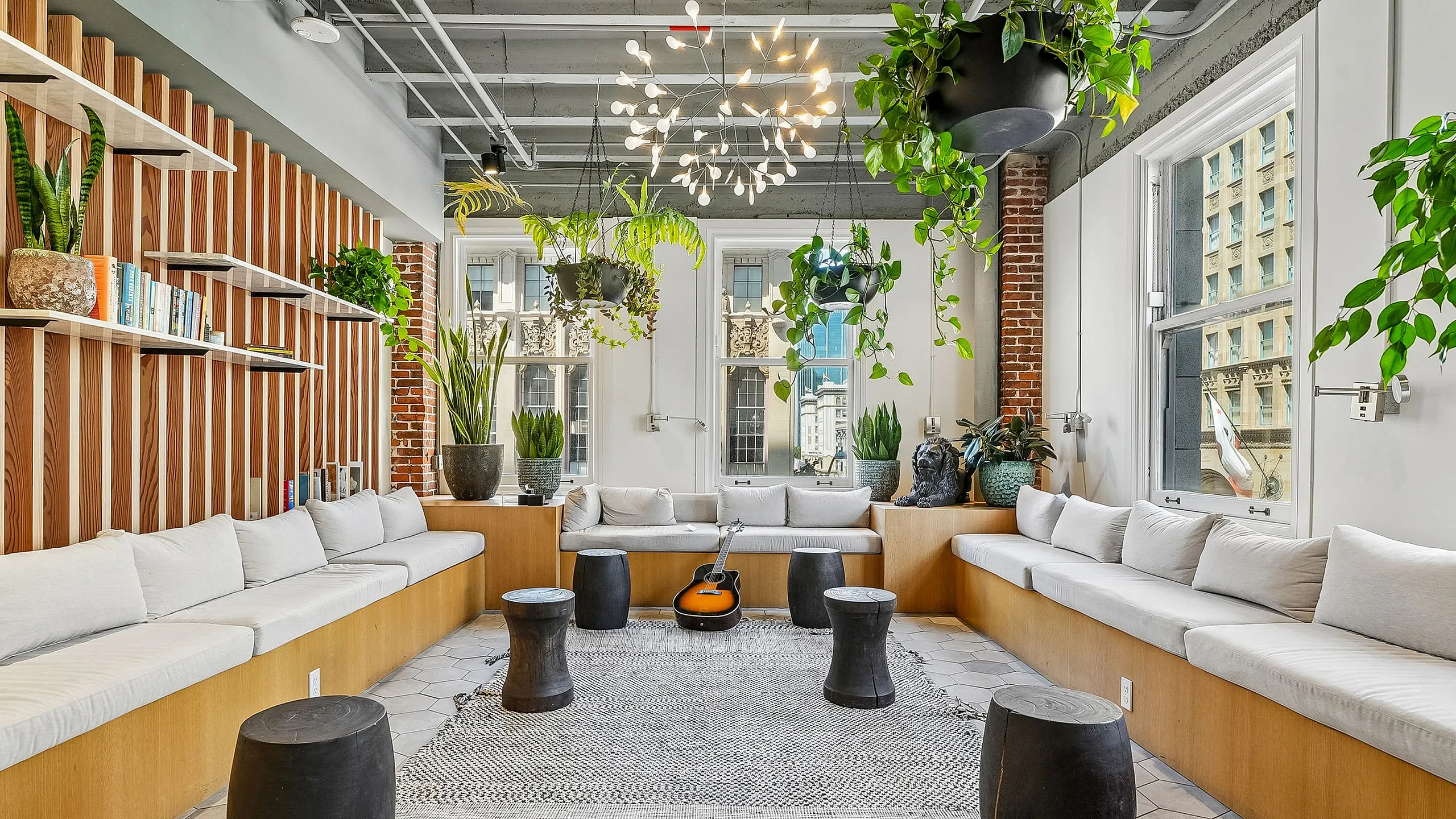 Modern communal living area with stylish seating, lush hanging plants, and a contemporary chandelier, offering a cozy ambiance with cityscape views.