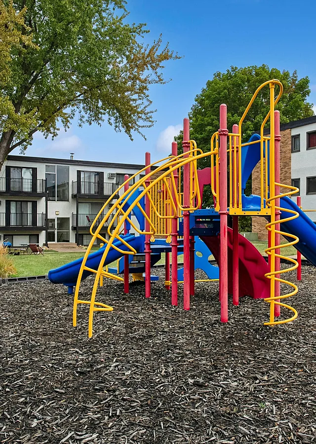 Inviting apartment complex with modern balconies, nestled next to a vibrant playground surrounded by lush greenery.