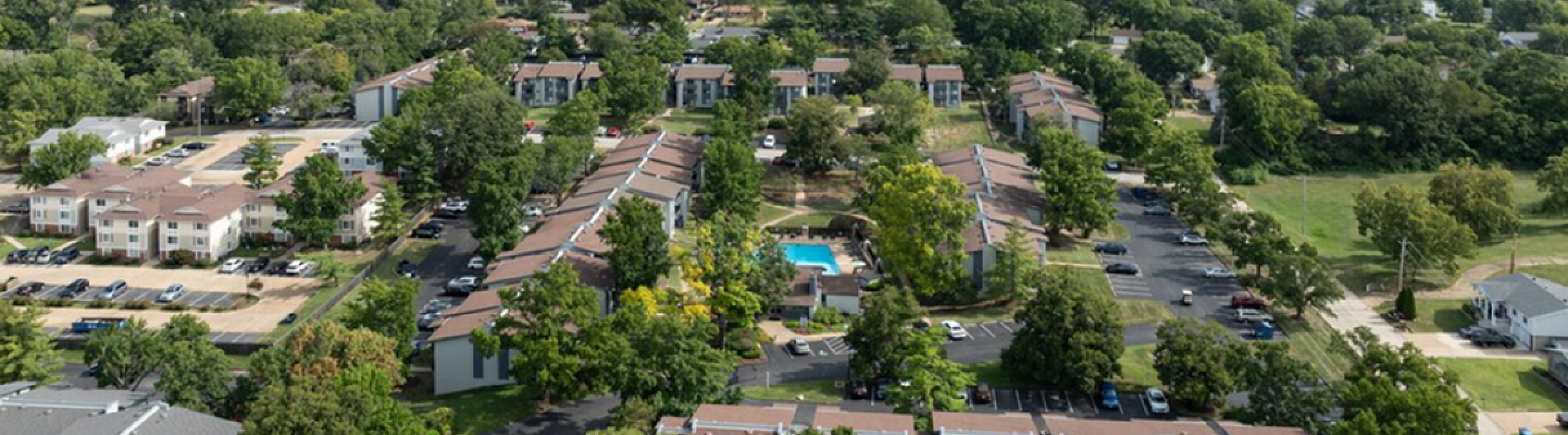 Aerial view of a lush apartment complex with multiple buildings, ample parking, and a sparkling central pool surrounded by vibrant greenery, offering a serene community living experience.