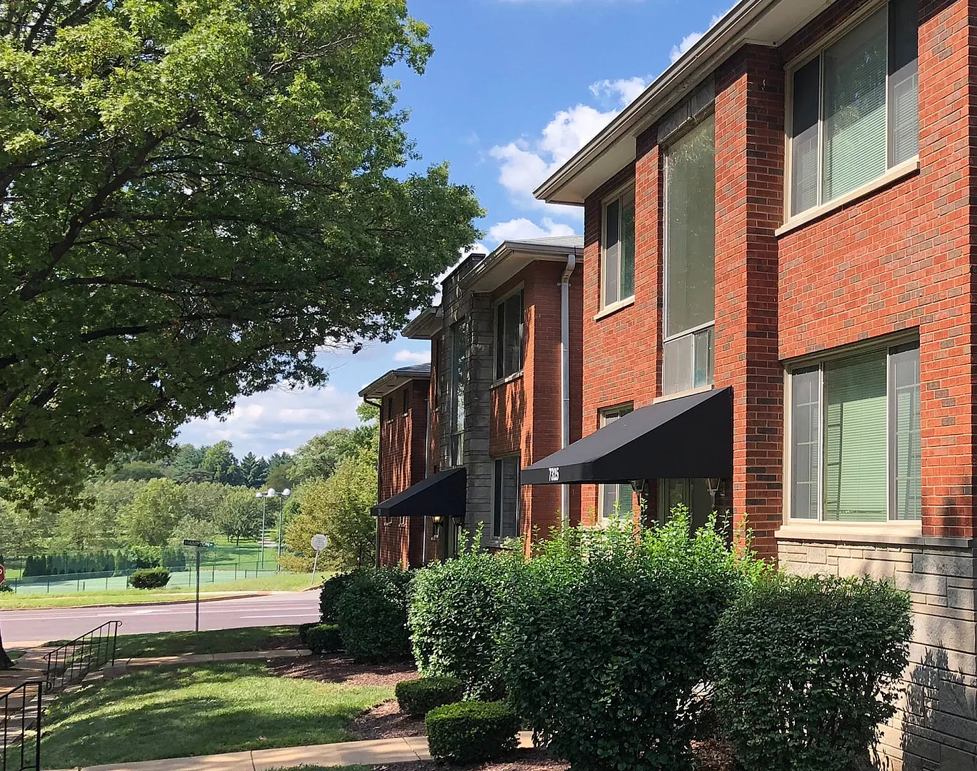 Charming brick apartment building surrounded by lush greenery on a sunny day, offering a peaceful residential vibe.