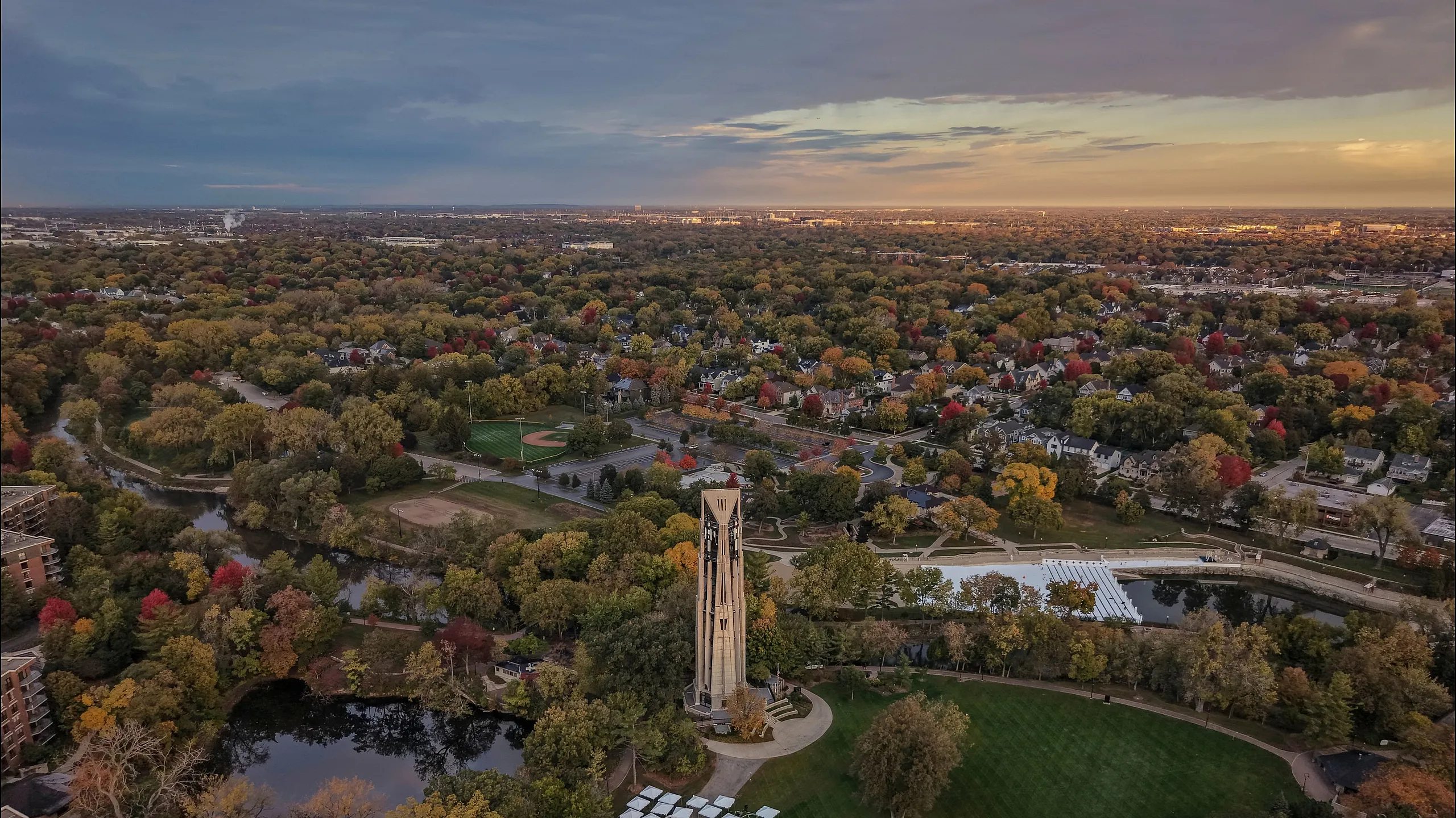 Breathtaking aerial view of a lush, expansive park with a central tower, surrounded by vibrant fall foliage and a serene pond.