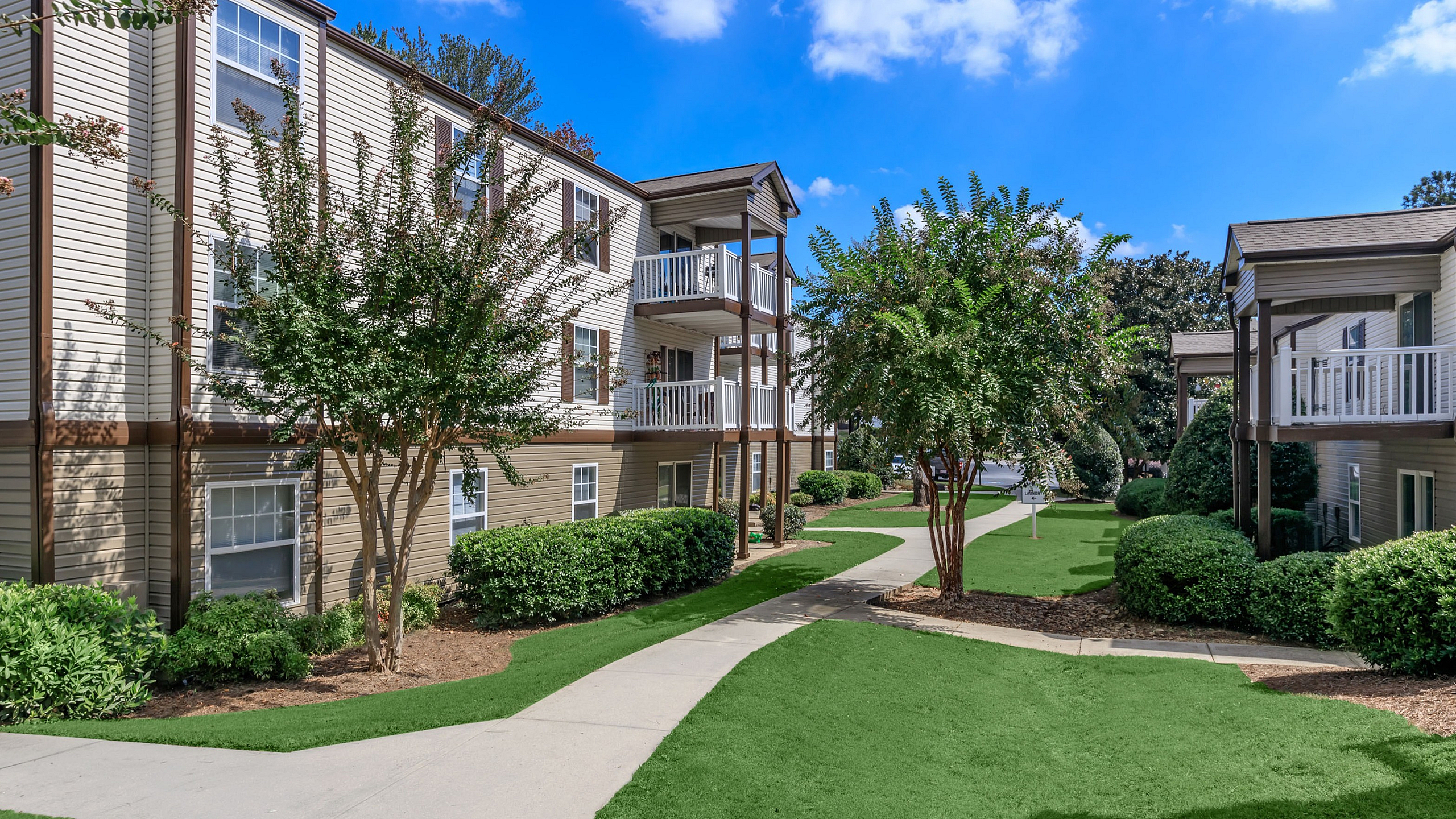 Charming apartment complex with lush green lawns, well-maintained pathways, and inviting balconies under a clear blue sky.