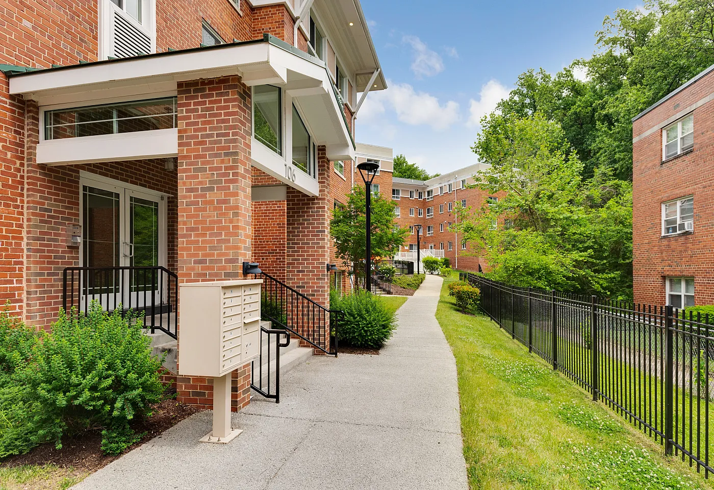 Charming brick apartment building with lush greenery and a welcoming entrance, featuring a picturesque walkway and modern mailboxes.